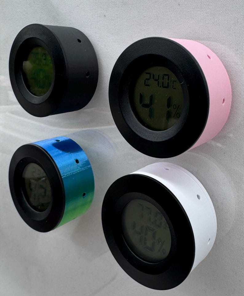 Puede incluir: Cuatro peque&ntilde;os higr&oacute;metros digitales redondos en negro, rosa, azul-verde y blanco. Cada uno tiene un bisel negro y una pantalla digital que muestra la temperatura y la humedad. Los higr&oacute;metros est&aacute;n montados sobre una superficie blanca.