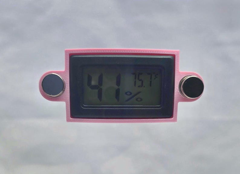 May include: A pink digital thermometer with a black display showing 41% humidity and 75.7&deg;F temperature. The device has two circular, dark gray buttons on either side.