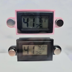 May include: Two digital hygrometers. The top one is pink, the bottom one is black. Both have a black display showing the temperature in Fahrenheit and the humidity percentage. Each has two silver-colored magnetic attachments.