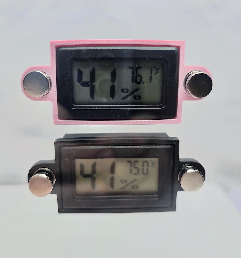 May include: Two digital hygrometers. The top one is pink, the bottom one is black. Both have a black display showing the temperature in Fahrenheit and the humidity percentage. Each has two silver-colored magnetic attachments.