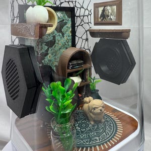 May include: A miniature diorama enclosed in a clear, curved glass case. Inside, there's a small dog figurine on a patterned rug, miniature furniture, and framed pictures. The scene includes a small bookshelf, plants, and decorative elements.