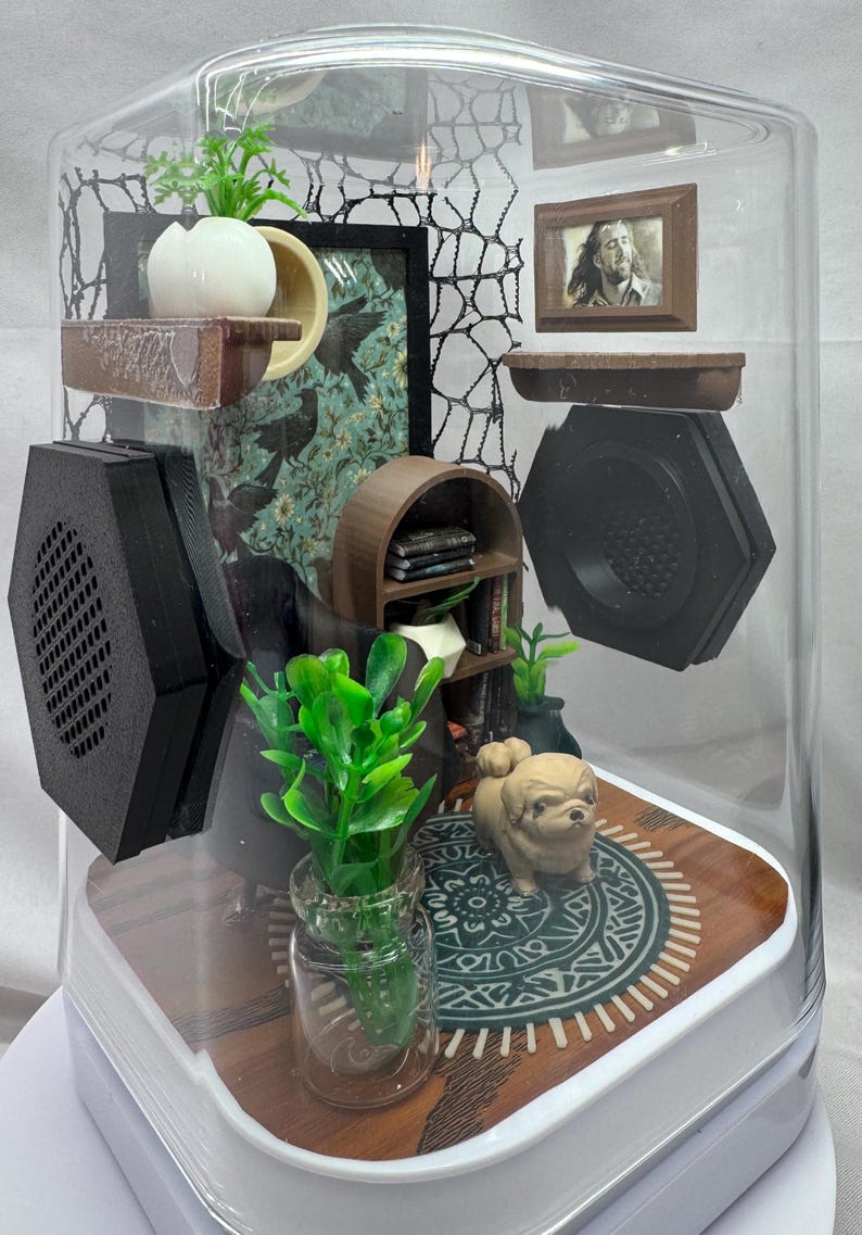 May include: A miniature diorama enclosed in a clear, curved glass case. Inside, there's a small dog figurine on a patterned rug, miniature furniture, and framed pictures. The scene includes a small bookshelf, plants, and decorative elements.