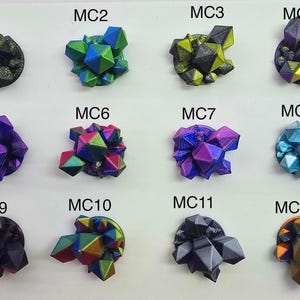May include: A collection of twelve colorful, geometric crystal sculptures. Each sculpture is a unique combination of colors, including green, blue, purple, and gold. The crystals are mounted on a white surface and labeled with the letters "MC" followed by a number.