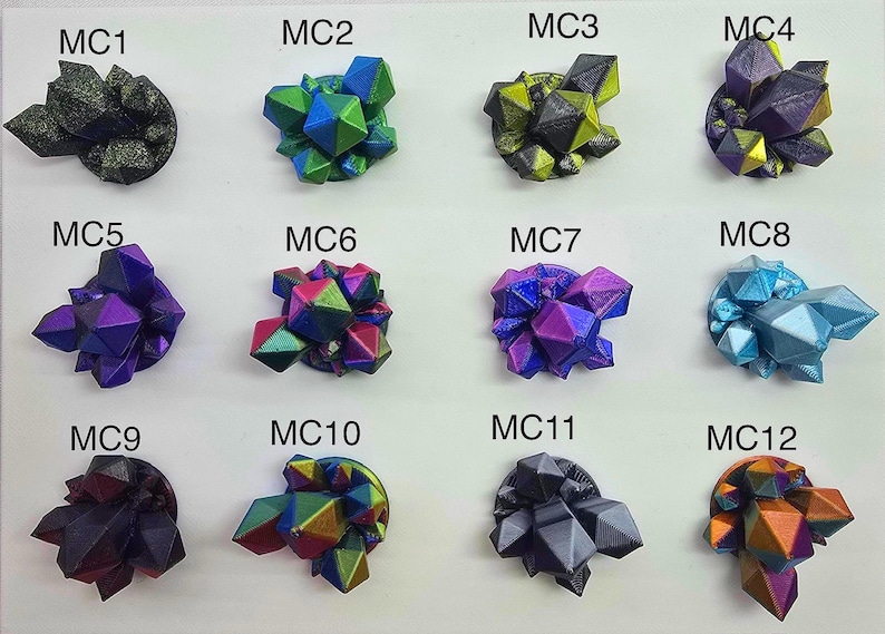 May include: A collection of twelve colorful, geometric crystal sculptures. Each sculpture is a unique combination of colors, including green, blue, purple, and gold. The crystals are mounted on a white surface and labeled with the letters "MC" followed by a number.