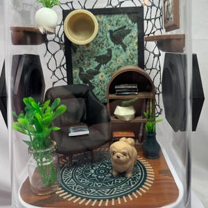 May include: Miniature diorama inside a clear dome. The scene includes a small black armchair, a bookshelf, a rug, and a decorative dog figurine. The background features a framed picture with bird imagery and a decorative net.