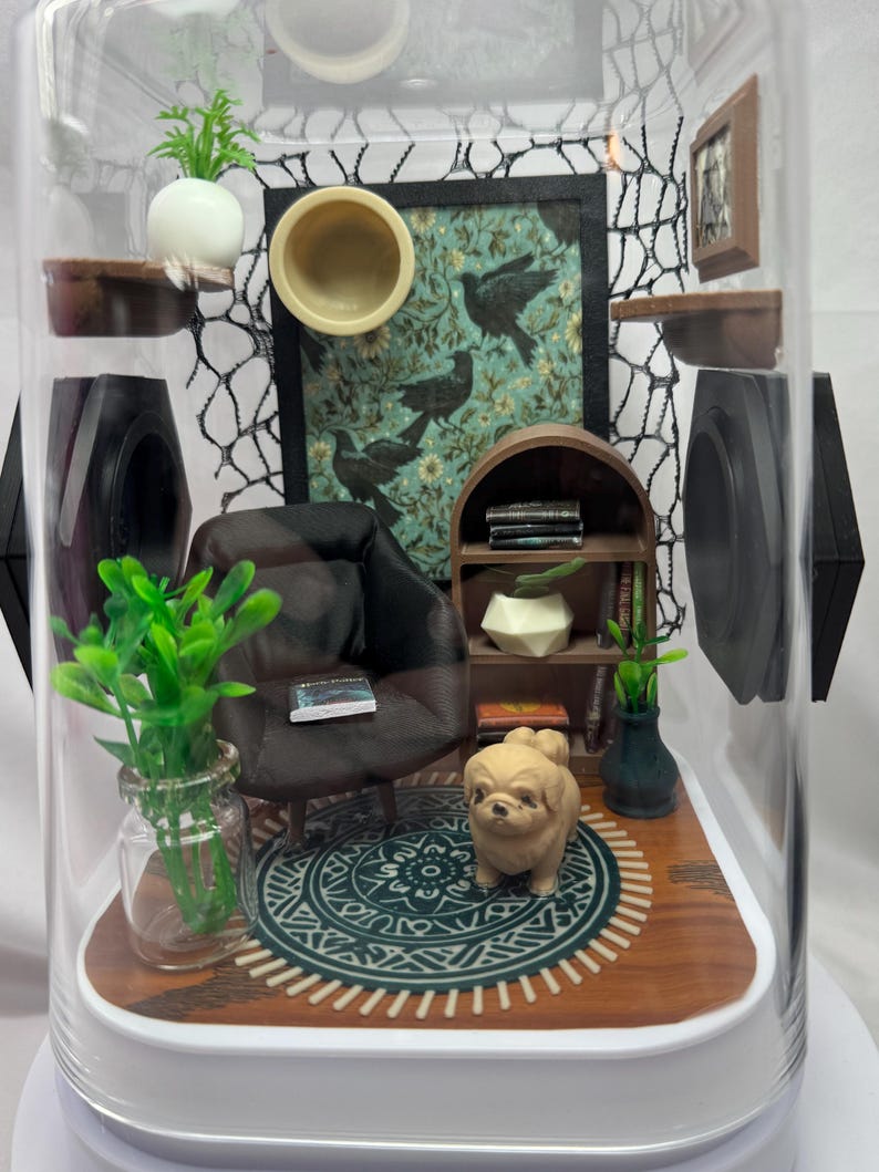 May include: Miniature diorama inside a clear dome. The scene includes a small black armchair, a bookshelf, a rug, and a decorative dog figurine. The background features a framed picture with bird imagery and a decorative net.