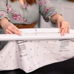 Peut inclure: Un tissu blanc avec des motifs de croix noirs est fixé à une barre blanche. Le tissu porte le texte "Grace" et "Premium Cloth Leaders". La personne porte une robe à motifs floraux.