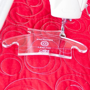Puede incluir: Una plantilla de acolchado de acrílico transparente con el texto "2-inch Feathers" y "Leah Day Made by Grace" se coloca sobre una tela acolchada roja. La plantilla está diseñada para ayudar con los patrones de plumas de acolchado.