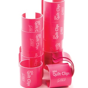 May include: Bright pink mini quilt clips stacked and arranged on a white background. The clips are cylindrical with a curved opening. The text "mini Quilt Clips" and "MADE BY Grace" are printed on the clips.