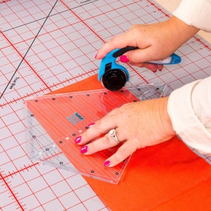 Puede incluir: Una persona usando un cortador rotatorio azul para cortar tela naranja sobre una alfombra de corte de cuadr&iacute;cula blanca y roja. La tela se est&aacute; cortando usando una regla de pl&aacute;stico transparente con la palabra "Tula Pink" impresa en ella.