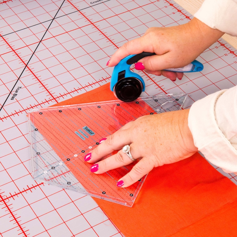 Puede incluir: Una persona usando un cortador rotatorio azul para cortar tela naranja sobre una alfombra de corte de cuadr&iacute;cula blanca y roja. La tela se est&aacute; cortando usando una regla de pl&aacute;stico transparente con la palabra "Tula Pink" impresa en ella.