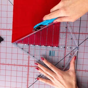 Pode incluir: Uma costureira usa um cortador rotativo azul e uma r&eacute;gua triangular transparente para cortar um peda&ccedil;o de tecido vermelho. A r&eacute;gua tem linhas de grade e marca&ccedil;&otilde;es de &acirc;ngulo. O corte &eacute; feito em uma base de corte cinza.