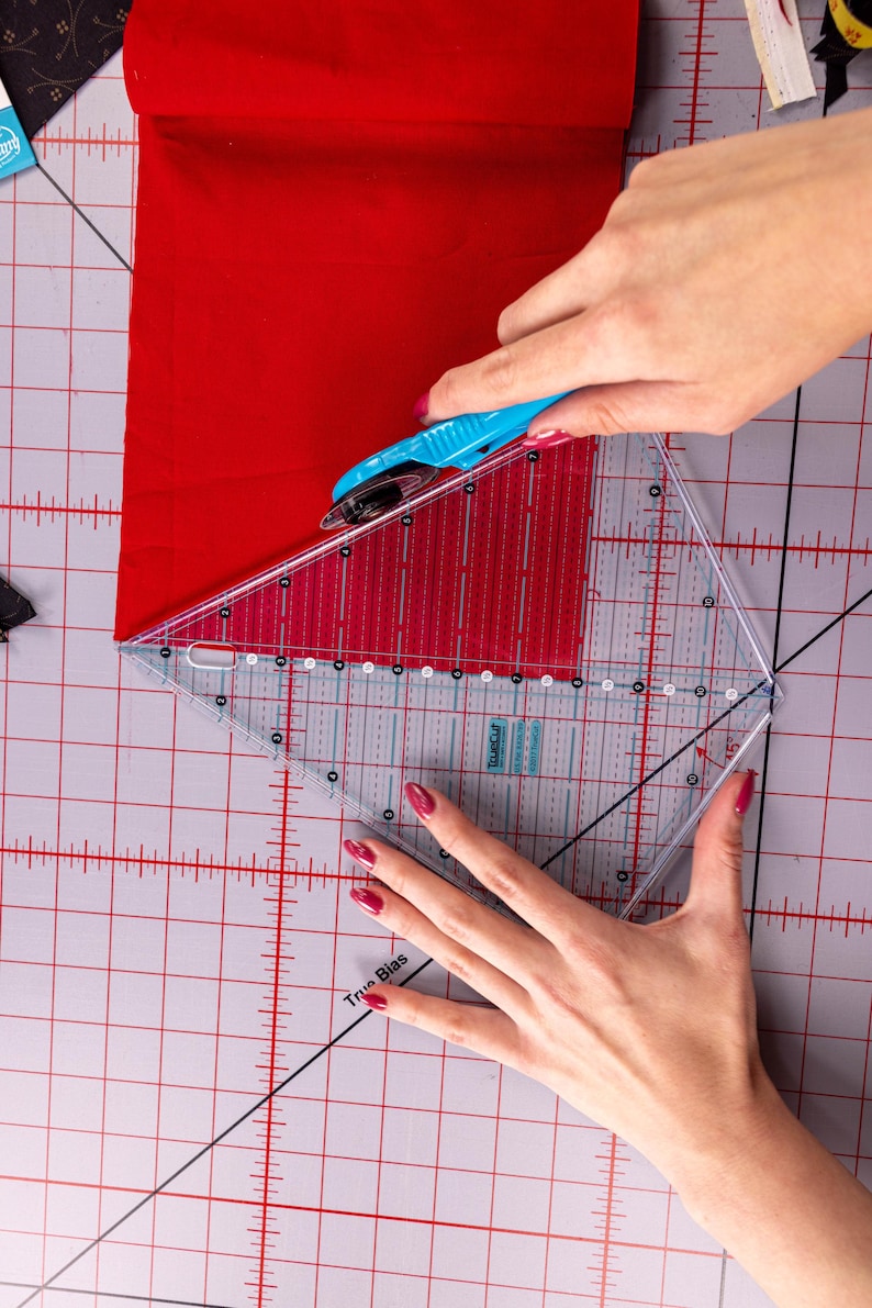 Pode incluir: Uma costureira usa um cortador rotativo azul e uma r&eacute;gua triangular transparente para cortar um peda&ccedil;o de tecido vermelho. A r&eacute;gua tem linhas de grade e marca&ccedil;&otilde;es de &acirc;ngulo. O corte &eacute; feito em uma base de corte cinza.