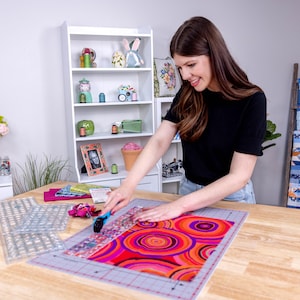 Pode incluir: Uma pessoa est&aacute; cortando tecido em uma mesa de madeira. O tecido tem um padr&atilde;o vibrante e colorido de c&iacute;rculos vermelhos, laranja e roxos. R&eacute;guas transparentes e uma base de corte tamb&eacute;m est&atilde;o sobre a mesa, sugerindo um projeto de costura ou patchwork.