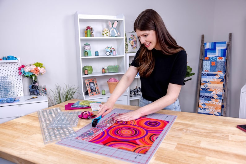 Pode incluir: Uma pessoa est&aacute; cortando tecido em uma mesa de madeira. O tecido tem um padr&atilde;o vibrante e colorido de c&iacute;rculos vermelhos, laranja e roxos. R&eacute;guas transparentes e uma base de corte tamb&eacute;m est&atilde;o sobre a mesa, sugerindo um projeto de costura ou patchwork.