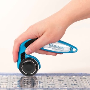 Puede incluir: Un cortador rotatorio azul y negro con un mango que dice "Truecut Easy Safe Accurate" se est&aacute; utilizando para cortar una regla de pl&aacute;stico transparente con un patr&oacute;n de cuadr&iacute;cula.