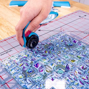 Pode incluir: Um cortador rotativo azul com uma l&acirc;mina preta corta tecido sobre uma base de corte. O tecido tem um padr&atilde;o floral em tons de azul, roxo e verde. Uma r&eacute;gua transparente &eacute; usada como guia.