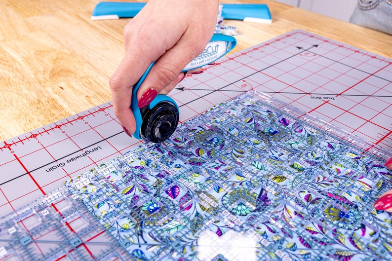 Pode incluir: Um cortador rotativo azul com uma l&acirc;mina preta corta tecido sobre uma base de corte. O tecido tem um padr&atilde;o floral em tons de azul, roxo e verde. Uma r&eacute;gua transparente &eacute; usada como guia.