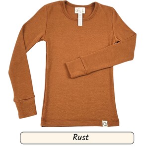 Könnte beinhalten: Ein langärmeliges rostfarbenes Shirt mit Rundhalsausschnitt und gerippten Bündchen. Das Shirt hat ein kleines Etikett am Ausschnitt und ein Etikett unten rechts. Das Wort "Rust" wird in einem rechteckigen Feld darunter angezeigt.