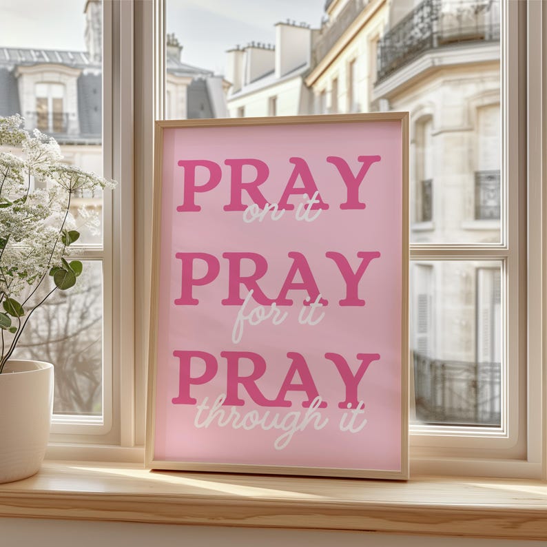 Może przedstawiać: Oprawiony r&oacute;żowy nadruk z napisami "PRAY on it, PRAY for it, PRAY through it" w r&oacute;żowych i białych czcionkach. Nadruk jest w jasnej drewnianej ramie, ustawionej przed oknem z widokiem na budynek w stylu europejskim.