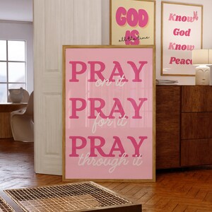 Może przedstawiać: Oprawiony r&oacute;żowy plakat z napisem "PRAY on it, PRAY for it, PRAY through it" w r&oacute;żowych literach. W tle widoczne są dwa inne oprawione plakaty z inspirującym tekstem. Pok&oacute;j ma drewniane podłogi.