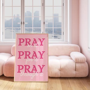 Może przedstawiać: Oprawiony r&oacute;żowy wydruk z napisami "PRAY on it, PRAY for it, PRAY through it" w r&oacute;żowej i białej czcionce. Wydruk znajduje się w jasno r&oacute;żowym pokoju z pasującą sofą i oknem z widokiem na panoramę miasta.