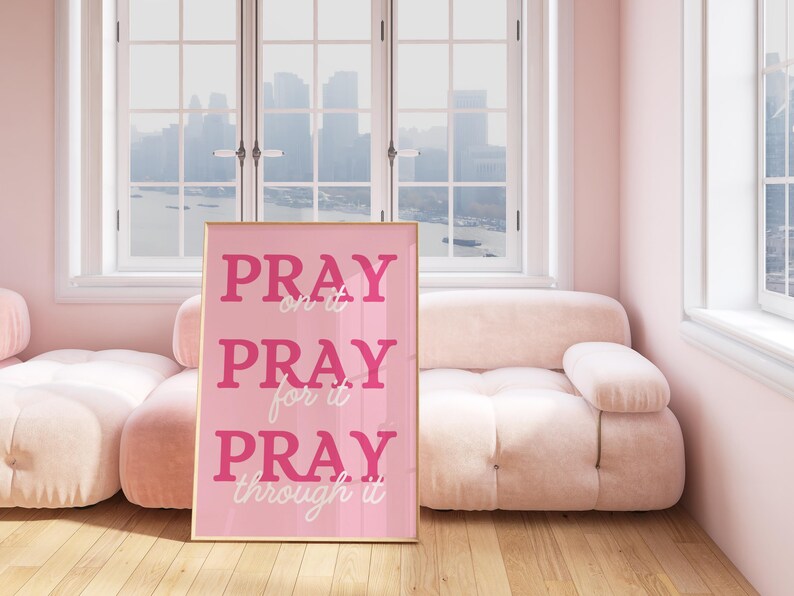Może przedstawiać: Oprawiony r&oacute;żowy wydruk z napisami "PRAY on it, PRAY for it, PRAY through it" w r&oacute;żowej i białej czcionce. Wydruk znajduje się w jasno r&oacute;żowym pokoju z pasującą sofą i oknem z widokiem na panoramę miasta.