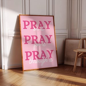Może przedstawiać: Oprawiony r&oacute;żowy plakat z napisem "PRAY on it, PRAY for it, PRAY through it" pogrubioną, r&oacute;żową czcionką. Plakat ma drewnianą ramę i opiera się o białą ścianę. Podłoga jest drewniana.