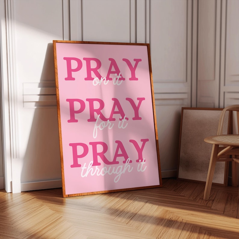 Może przedstawiać: Oprawiony r&oacute;żowy plakat z napisem "PRAY on it, PRAY for it, PRAY through it" pogrubioną, r&oacute;żową czcionką. Plakat ma drewnianą ramę i opiera się o białą ścianę. Podłoga jest drewniana.