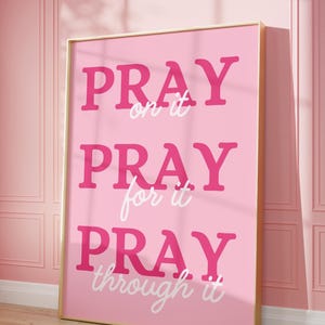 Może przedstawiać: Oprawiony r&oacute;żowy wydruk artystyczny ze słowami "PRAY on it, PRAY for it, PRAY through it" pogrubioną, r&oacute;żową czcionką. Grafika jest umieszczona na r&oacute;żowym tle i oprawiona w złoto. Wydruk opiera się o r&oacute;żową ścianę.