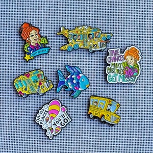Op de afbeelding: Een verzameling van zeven kleurrijke emaille pins met stripfiguren, een gele schoolbus, een vliegende bus, een blauwe vis en teksten waaronder "DO IT FOR HER," "TAKE CHANCES MAKE MISTAKES GET MESSY" en "OH! the PLACES You'll GO!"
