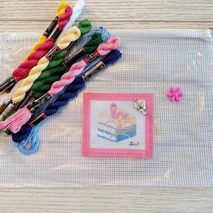 May include: A clear mesh zip-up pouch containing several skeins of embroidery thread in yellow, pink, green, blue, and white. A small square needlepoint canvas with a pink border features a stitched pink worm on books, with a needle and a pink flower.