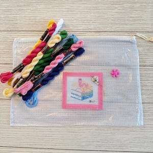 May include: A cross-stitch kit featuring a selection of colourful embroidery threads, a pink-framed cross-stitch, a pink flower, and a clear mesh bag. The cross-stitch design depicts a cake with a flamingo. The threads are in shades of pink, yellow, green, and blue.