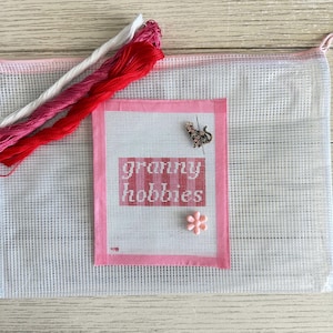 Op de afbeelding: Een kruissteekpakket met de woorden "granny hobbies" in rood en wit. De set bevat roze en rode borduurgaren, een mesh tas, een katvormige speld en een roze bloemvormige speld.