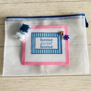 May include: A needlepoint kit with a canvas that reads "home sweet home" in blue and white. The canvas is framed in pink and includes a small bee charm and a purple flower pin. The kit is inside a white mesh zipper pouch with a blue zipper.