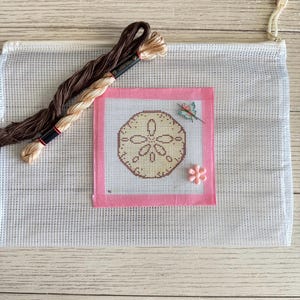 May include: A needlepoint kit with a sand dollar design in beige and brown, framed by a pink border. Includes brown and beige embroidery thread, a butterfly pin, and a pink flower pin. The kit is set against a white mesh background.