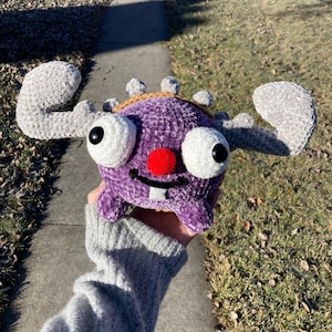 May include: A purple crocheted plush toy with large white eyes, a red nose, and a single white tooth. The toy has grey crocheted antlers and is held in a hand.