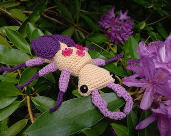 May include: A handmade crochet insect toy in shades of beige, purple, and pink, resting on green leaves with purple flowers in the background. The toy has a textured body, six legs, and large black eyes.