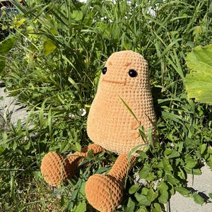 May include: A light brown, handmade crochet potato plush toy. The toy has a smiling face with black eyes and small arms and legs. It is sitting amongst green grass and weeds. The toy is made of soft yarn.
