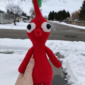 May include: A red crocheted plush toy with a green leaf on top and large white eyes. The toy is being held in a person's hand.