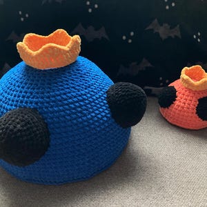 May include: Two crocheted stuffed toys, one blue with a yellow crown and black eyes, the other orange with a yellow crown and black spots.