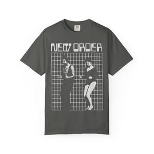 Puede incluir: Una camiseta gris carbón de manga corta con un estampado gráfico blanco de la banda New Order. El diseño incluye el texto NEW ORDER sobre un patrón de cuadrícula con dos figuras bailando, representado en un estilo de tinta blanca de alto contraste.