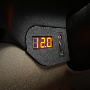 May include: A black digital display with the number 12.0 in orange lights. The display is mounted in a black plastic housing with a small black toggle switch.