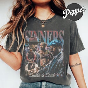 May include: A charcoal gray short-sleeve t-shirt featuring a vintage-style graphic print with the text "SINNERS" and "Smoke & Stack". The design includes multiple photographic portraits of a man in various outfits, accented by star motifs.