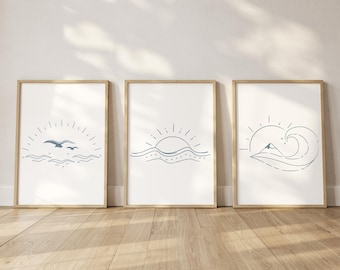 May include: Three framed art prints with minimalist line drawings. The prints feature a sunrise over waves, a sun with birds, and a mountain with a wave. The frames are a light wood color, and the art is in shades of blue on a white background.