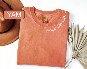 May include: A coral colored t-shirt with the word "mama" embroidered in white on the neckline. The shirt is folded and laid flat on a white surface.