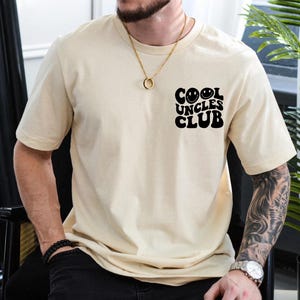 Può includere: Una maglietta beige con una grafica nera che dice "Cool Uncles Club" con due faccine sorridenti.