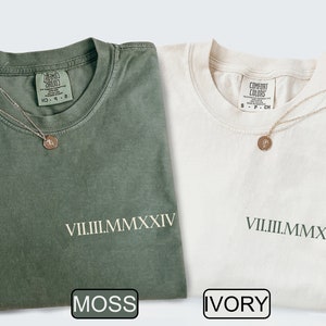 May include: Two t-shirts, one moss green and one ivory, are displayed. Each shirt has the roman numeral date VII.III.MMXXIV printed on the front. A delicate gold necklace with a pendant rests on each shirt. The shirts are labeled "MOSS" and "IVORY".