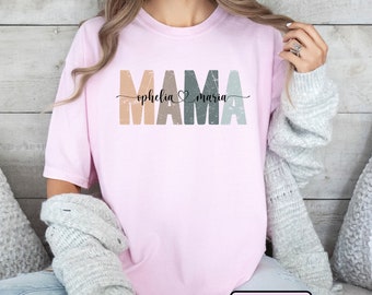 May include: Pink t-shirt with the word "MAMA" in colorful distressed letters. The names "ophelia" and "maria" are written in cursive script with a heart symbol between them.