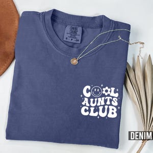 Può includere: Una maglietta blu navy con una grafica bianca che dice "Cool Aunts Club" con un viso sorridente e un fiore.
